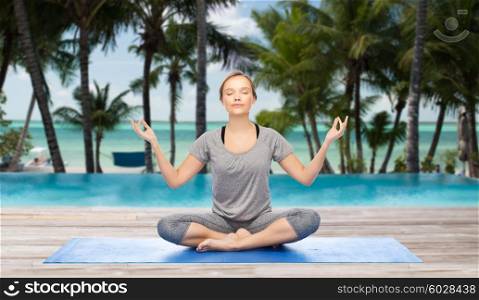 fitness, sport, people and healthy lifestyle concept - woman making yoga meditation in lotus pose on mat over hotel resort pool on tropical beach background