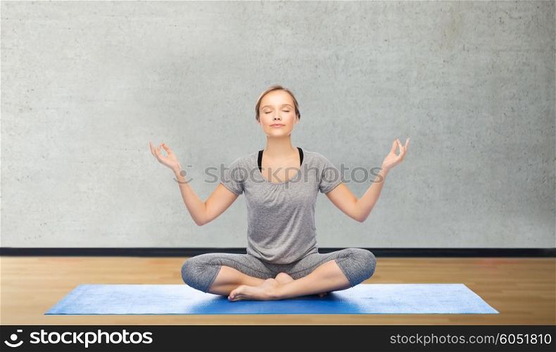 fitness, sport, people and healthy lifestyle concept - woman making yoga meditation in lotus pose on mat over gym room background
