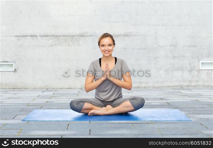 fitness, sport, people and healthy lifestyle concept - woman making yoga meditation in lotus pose on mat over urban street background