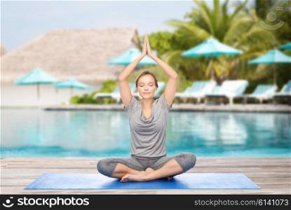 fitness, sport, people and healthy lifestyle concept - woman making yoga meditation in lotus pose on mat over beach and swimming pool background