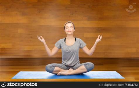 fitness, sport, people and healthy lifestyle concept - woman making yoga meditation in lotus pose on mat over wooden room background