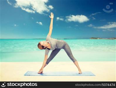 fitness, sport, people and healthy lifestyle concept - woman making yoga triangle pose on mat over beach background. woman making yoga triangle pose on mat