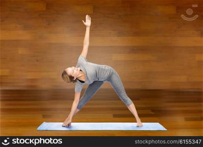 fitness, sport, people and healthy lifestyle concept - woman making yoga triangle pose on mat over wooden room background
