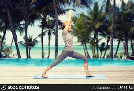 fitness, sport, people and healthy lifestyle concept - woman making yoga warrior pose on mat over hotel resort pool on tropical beach background. woman making yoga warrior pose on mat