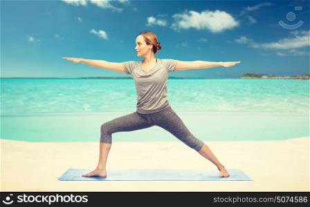 fitness, sport, people and healthy lifestyle concept - woman making yoga warrior pose on mat over sea and sky background. woman making yoga warrior pose on mat