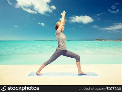 fitness, sport, people and healthy lifestyle concept - woman making yoga warrior pose on mat over beach background. woman making yoga warrior pose on mat