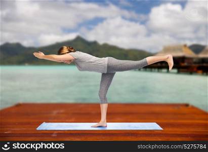 fitness, sport, people and healthy lifestyle concept - woman making yoga warrior pose on wooden pier over island beach and bungalow background. woman making yoga warrior pose on mat outdoors