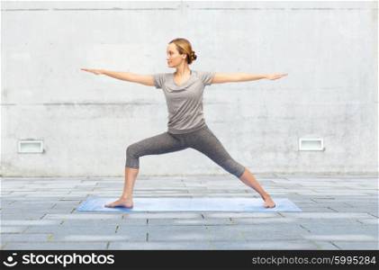fitness, sport, people and healthy lifestyle concept - woman making yoga warrior pose on mat over urban street background