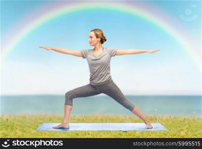 fitness, sport, people and healthy lifestyle concept - woman making yoga warrior pose on mat over blue sky, rainbow and sea background