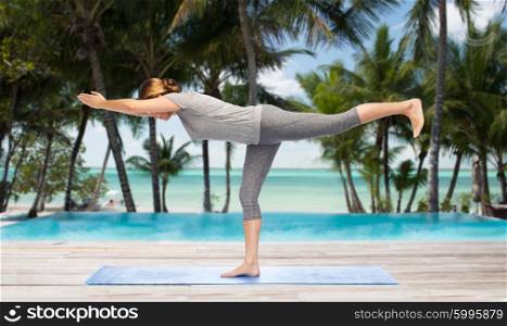 fitness, sport, people and healthy lifestyle concept - woman making yoga warrior pose on mat over hotel resort pool on tropical beach background