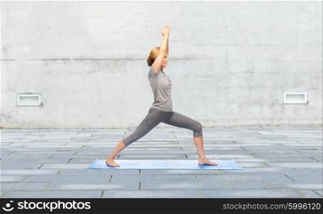 fitness, sport, people and healthy lifestyle concept - woman making yoga warrior pose on mat over urban street background