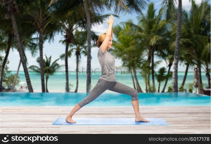 fitness, sport, people and healthy lifestyle concept - woman making yoga warrior pose on mat over hotel resort pool on tropical beach background