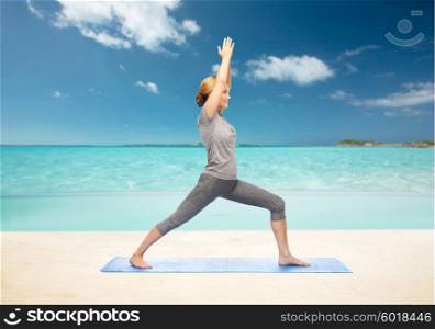 fitness, sport, people and healthy lifestyle concept - woman making yoga warrior pose on mat over beach background