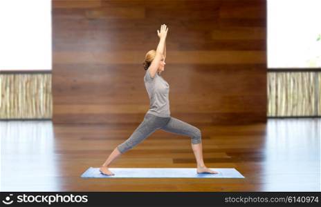 fitness, sport, people and healthy lifestyle concept - woman making yoga warrior pose on mat over wooden gym background