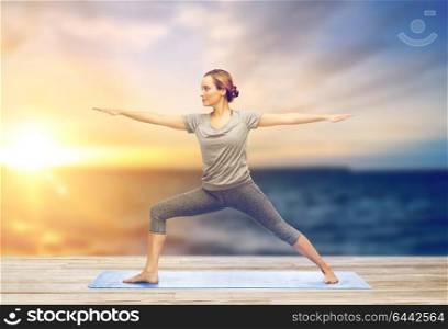 fitness, sport, people and healthy lifestyle concept - woman making yoga warrior pose on pier over sea background. woman making yoga warrior pose on mat