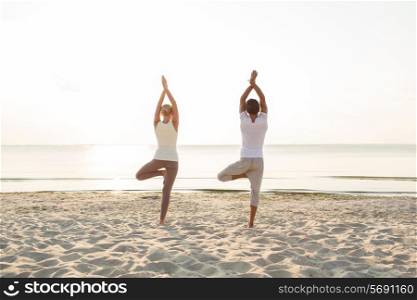 fitness, sport, people and lifestyle concept - couple making yoga exercises on sand outdoors from back