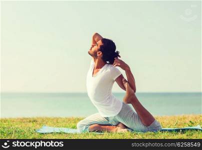 fitness, sport, people and lifestyle concept - smiling man making yoga exercises sitting on mat outdoors