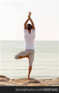 fitness, sport, people and lifestyle concept - young man making yoga exercises on beach from back