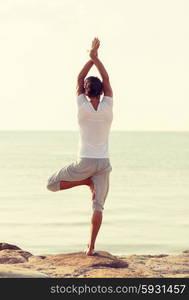 fitness, sport, people and lifestyle concept - young man making yoga exercises on beach from back