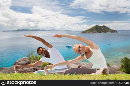 fitness, sport, people and recreation concept - smiling couple making yoga exercises sitting on mats outdoors over ocean background. smiling couple making yoga exercises outdoors