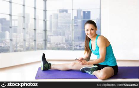 fitness, sport, people and technology concept - smiling woman with smartphone sitting on exercise mat over gym background