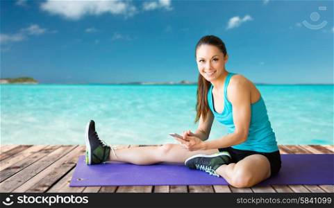 fitness, sport, people and technology concept - smiling woman with smartphone sitting on exercise mat over sea and wooden berth at resort background