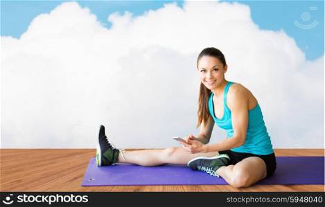fitness, sport, people and technology concept - smiling woman with smartphone sitting on exercise mat over wooden floor and sky with white cloud background