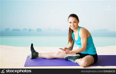 fitness, sport, people and technology concept - smiling woman with smartphone sitting on exercise mat over sea and pool at hotel resort background