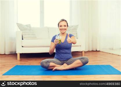 fitness, sport, people, diet and healthy lifestyle concept - happy woman with cup of smoothie sitting on mat at home and showing thumbs up. woman with smoothie showing thumbs up at home
