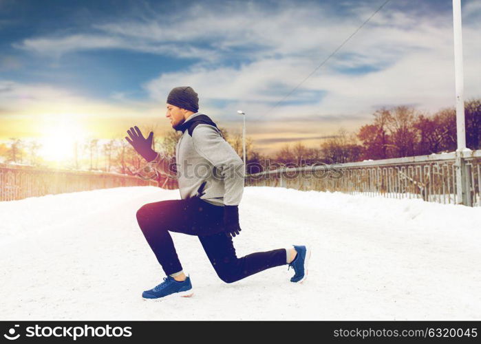 fitness, sport, people, exercising and healthy lifestyle concept - young man doing squats and warming up on snow covered winter bridge. man exercising and doing squats on winter bridge