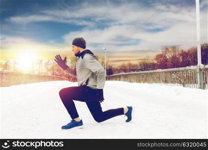 fitness, sport, people, exercising and healthy lifestyle concept - young man doing squats and warming up on snow covered winter bridge. man exercising and doing squats on winter bridge