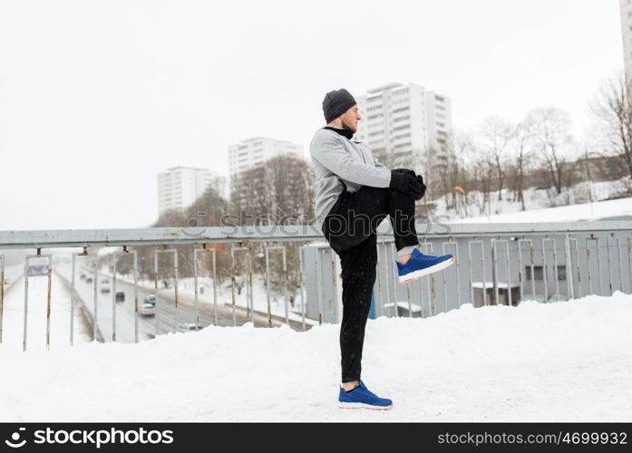 fitness, sport, people, exercising and healthy lifestyle concept - young man stretching leg and warmig up on snow covered winter bridge