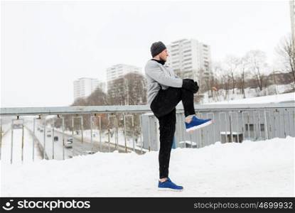 fitness, sport, people, exercising and healthy lifestyle concept - young man stretching leg and warmig up on snow covered winter bridge