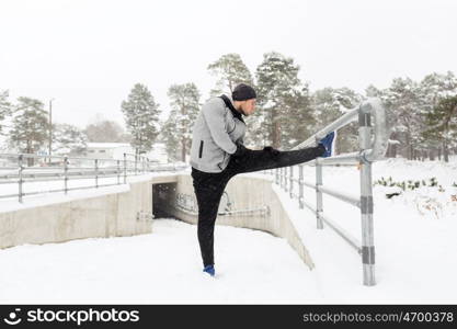 fitness, sport, people, exercising and healthy lifestyle concept - young man stretching leg and warming up at fence in winter