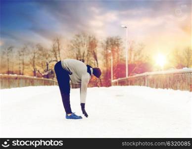 fitness, sport, people, exercising and healthy lifestyle concept - young man stretching leg and warming up on snow covered winter bridge. man exercising and stretching leg on winter bridge