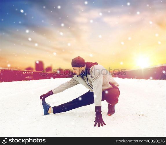 fitness, sport, people, exercising and healthy lifestyle concept - young man stretching leg and warmig up on snow covered winter bridge. man exercising and stretching leg on winter bridge