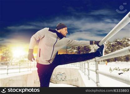 fitness, sport, people, exercising and healthy lifestyle concept - young man stretching leg and warming up at fence in winter. sports man stretching leg at fence in winter