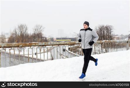 fitness, sport, people, music and healthy lifestyle concept - young man in earphones running along snow covered winter bridge