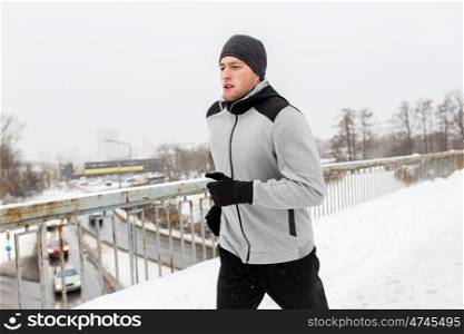 fitness, sport, people, music and healthy lifestyle concept - young man in earphones running along snow covered winter bridge