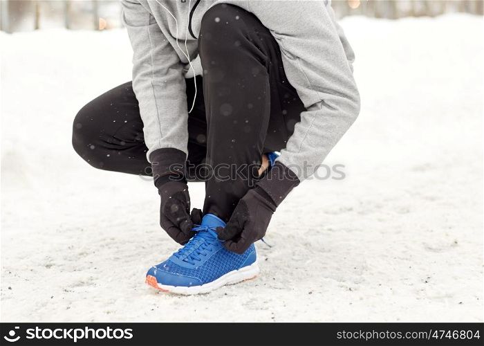 fitness, sport, people, music and healthy lifestyle concept - young man in earphones tying shoe on winter bridge