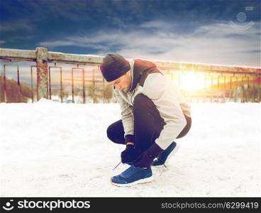 fitness, sport, people, music and healthy lifestyle concept - young man in earphones tying shoe on winter bridge. man with earphones tying sports shoes in winter