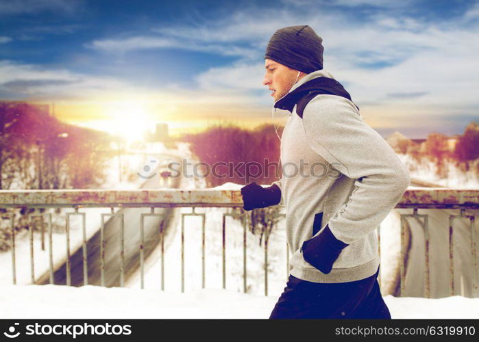 fitness, sport, people, music and healthy lifestyle concept - young man in earphones running along snow covered winter bridge. man in earphones running along winter bridge