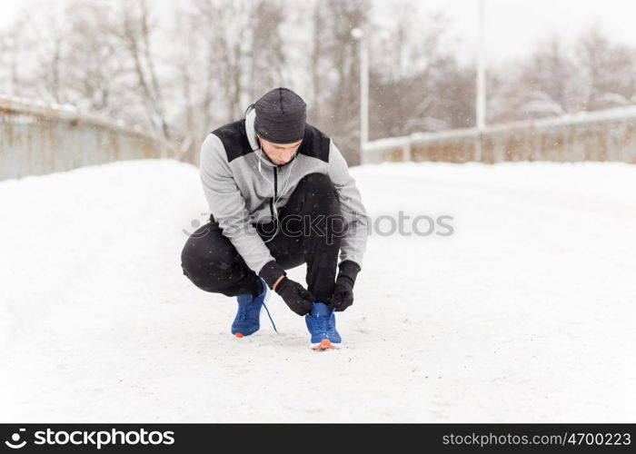 fitness, sport, people, music and healthy lifestyle concept - young man man in earphones tying shoe on winter bridge