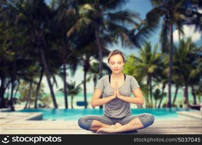 fitness, sport, people, resort and healthy lifestyle concept - woman making yoga meditation in lotus pose over swimming pool and tropical beach with palm trees background. woman making yoga meditation in lotus pose