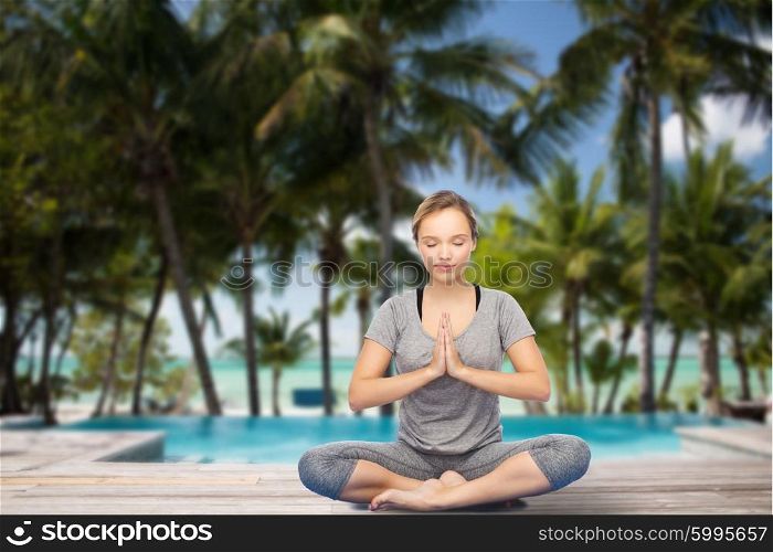 fitness, sport, people, resort and healthy lifestyle concept - woman making yoga meditation in lotus pose over swimming pool and tropical beach with palm trees background