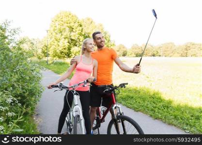 fitness, sport, people, technology and healthy lifestyle concept - happy couple with bicycle taking picture by smartphone on selfie stick outdoors