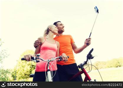 fitness, sport, people, technology and healthy lifestyle concept - happy couple with bicycle taking picture by smartphone on selfie stick outdoors