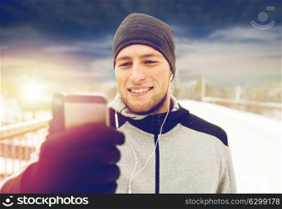 fitness, sport, people, technology and healthy lifestyle concept - happy smiling young man in earphones with smartphone listening to music on winter bridge. happy man with earphones and smartphone in winter