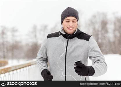 fitness, sport, people, technology and healthy lifestyle concept - happy young man in earphones with smartphone listening to music and running along winter road