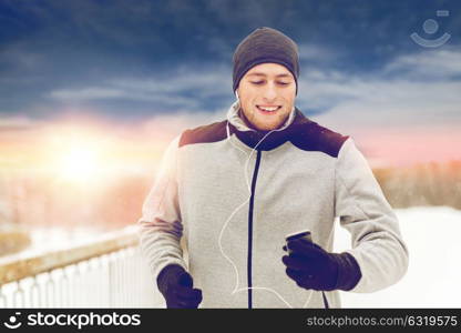 fitness, sport, people, technology and healthy lifestyle concept - happy young man in earphones with smartphone listening to music and running along winter road. happy man with earphones and smartphone in winter